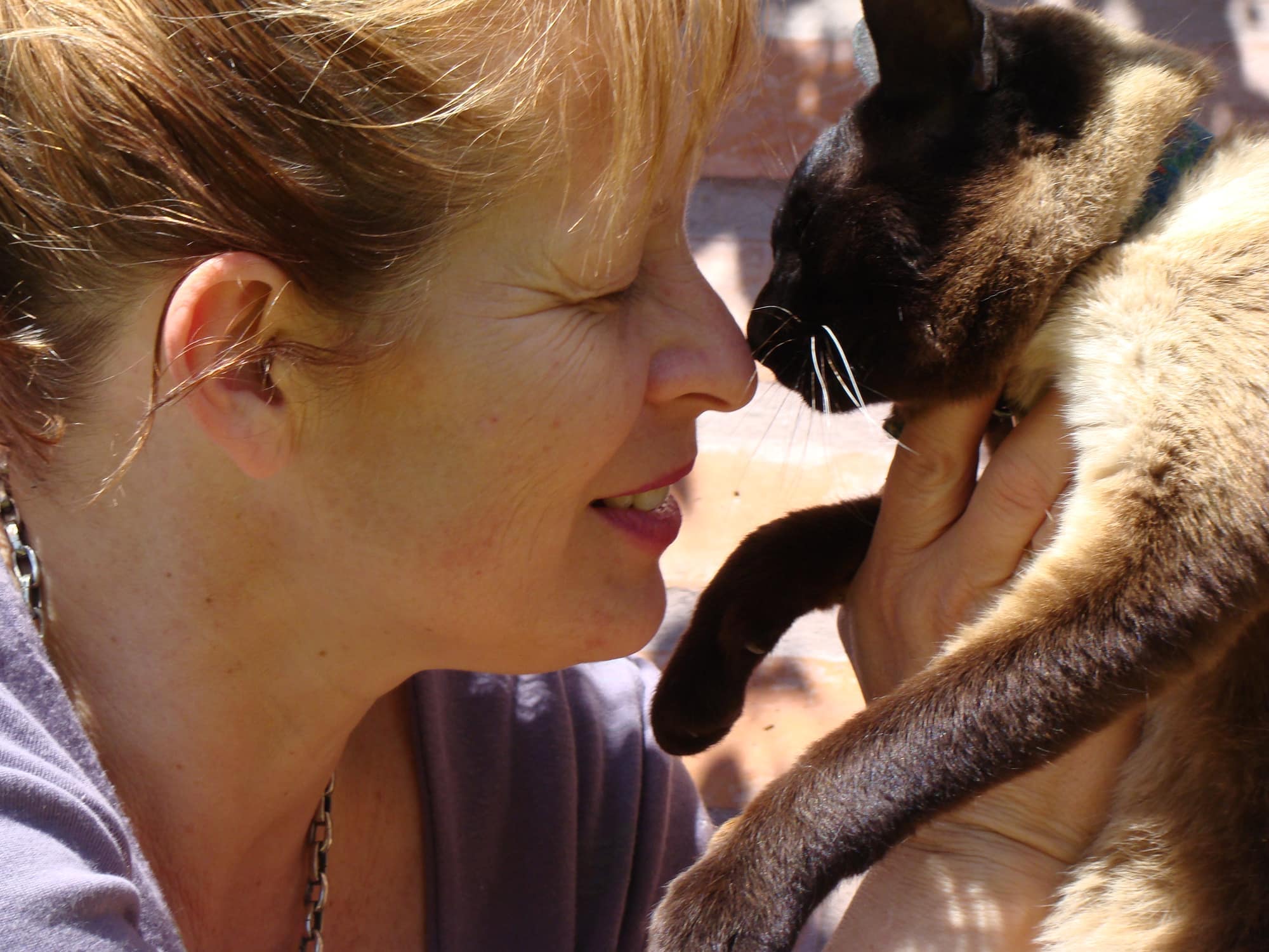 Una mujer sonríe con los ojos cerrados y junta su nariz con la de su gato siamés, que sostiene entre las manos bajo la luz cálida del sol.